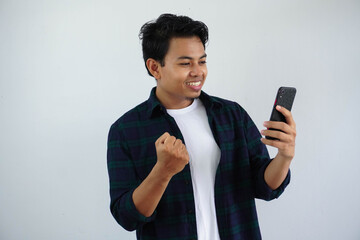 young asian man clenching fist showing excitement when looking to his phone isolated on white background - obrazy, fototapety, plakaty