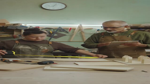 Vertical shot of experienced male carpenter with tablet entering sizes of wooden workpieces into database while his young female apprentice with Down syndrome taking measures of details