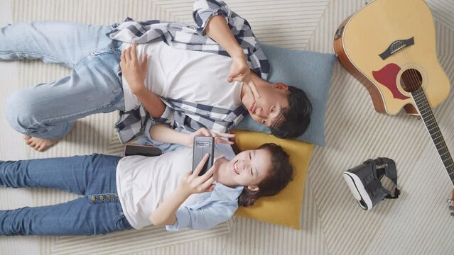 Top View Of Asian Teen Couple Using Smartphone Taking Selfie While Lying On Carpet On The Floor At Home. Smiling, Showing Peace And Heart Gesture
