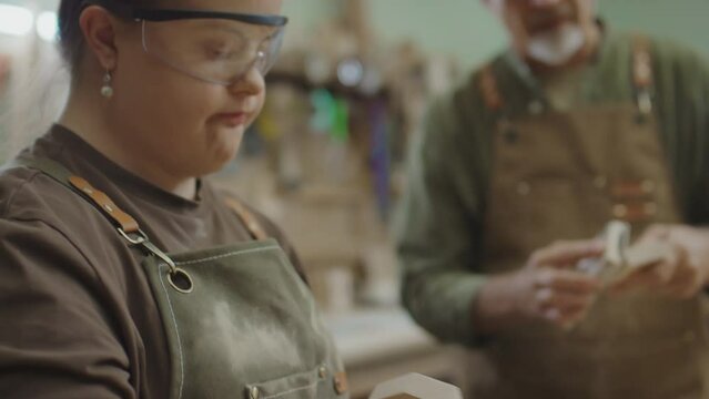 Tilt up of young woman with Down syndrome in apron and protective eyeglasses polishing wooden detail with handtool while standing in front of camera against mature woodworker