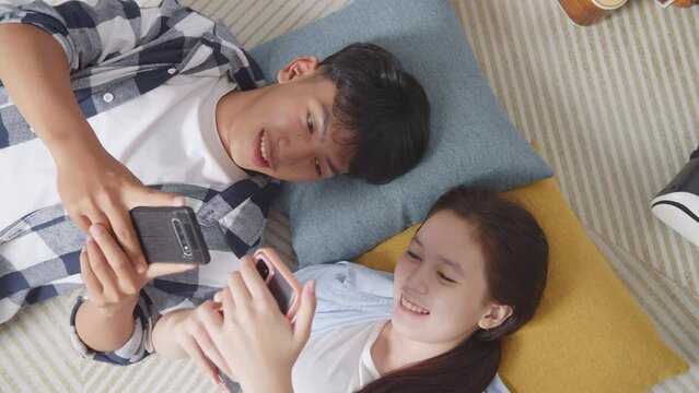 Close Up Top View Of Asian Teen Couple Playing Smartphone While Lying On Carpet On The Floor At Home. Speaking, And Enjoying Time Together
