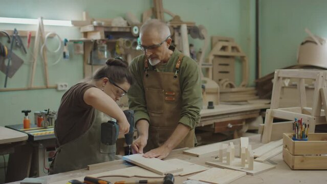 Mature joiner in apron explaining his apprentice with disability how to use electric drill and helping her during work with wooden workpieces while both standing by workbench