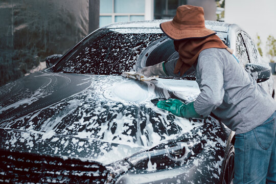 Woman worker washing car at car station outdoors with foam and yellow sponge.