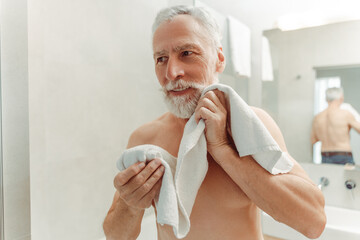 Handsome smiling senior man wiping face, standing in bathroom, holding towel, looking away