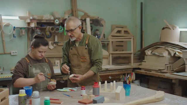 Young woman with Down syndrome and mature carpenter in workwear painting with gouache small wooden details for restoration of furniture while standing by workbench with supplies