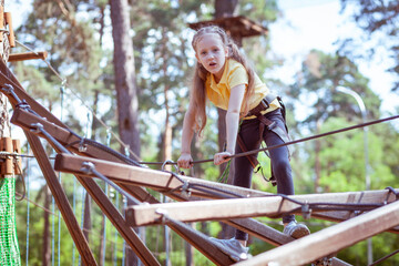 A child in a forest adventure park made of ropes. The girl is climbing the path with a high rope. Children's outdoor climbing entertainment center. Playground for children and sports with a cable car.