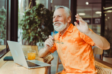 Smiling handsome senior man wearing stylish orange shirt holding eyeglasses using laptop waving hand