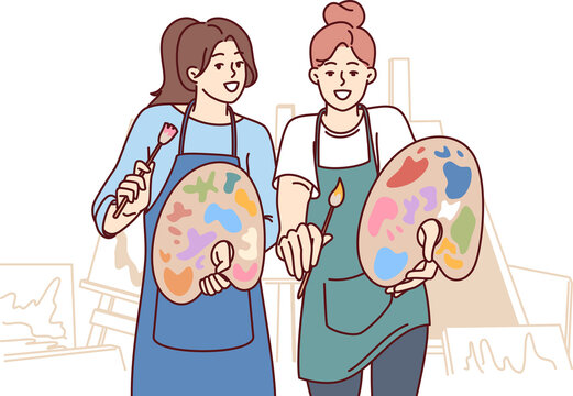 Two women artists hold es and palettes standing in workshop with easels to create artworks