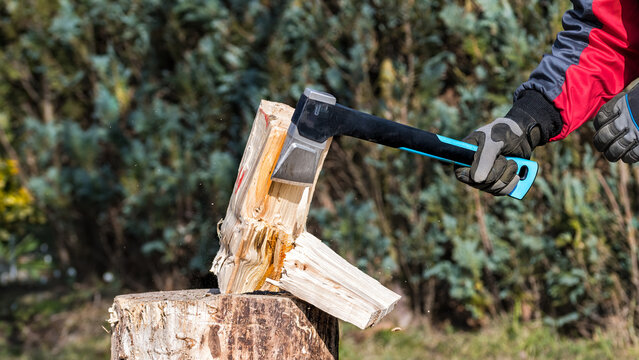 Closeup of sharp axe in human hand when splitting wood log on chopping block in garden. Working man holding modern cutting tool with metal blade and plastic haft. Firewood processing in energy crisis.