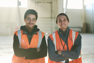 Two happy young multietnic foremen or workers of warehouse in safety jackets standing in front of camera with sunlight on background - obrazy, fototapety, plakaty