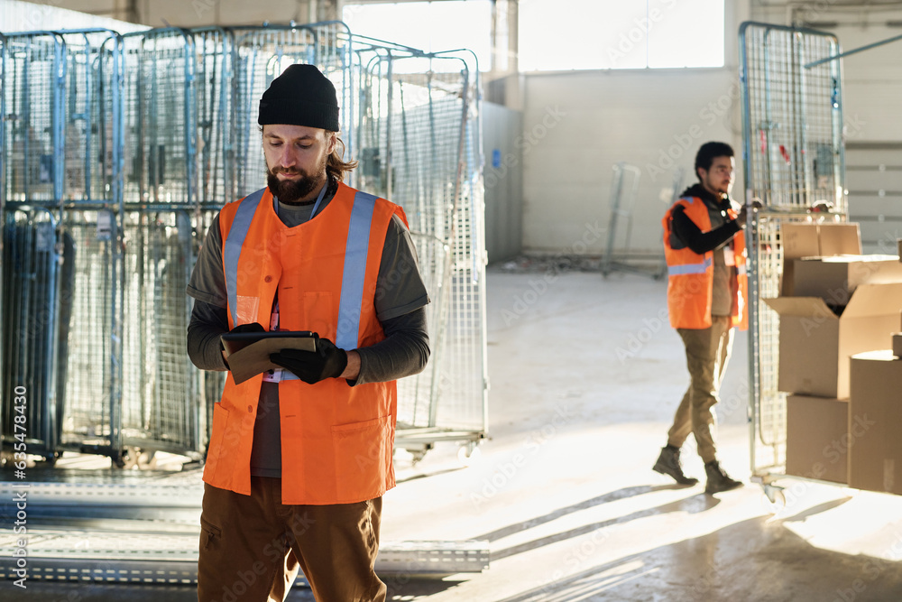 Leinwandbilder young worker of warehouse in protective reflective vest using tablet while standing in front of came