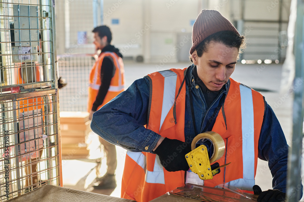 Leinwandbilder young man in gloves and workwear sealing packages with cellotape while standing in front of camera a