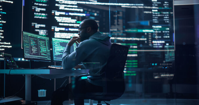 Portrait of a Black Man Working on Computer, Typing Lines of Code that Appear on Big Screens Surrounding him in Monitoring Room. Male Programmer Creating Innovative Software Using AI Data and System