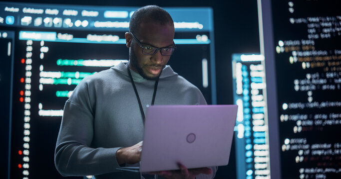 Portrait of Professional Programmer Developing a Big Data Interface Software Project.Young Black Man Working on Laptop Computer, Looking at Big Digital Screen Displaying Back-end Code Lines - Powered by Adobe
