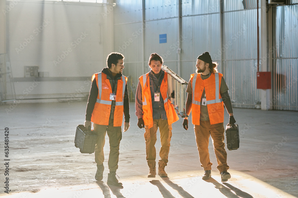 Poster three young intercultural engineers in workwear moving along warehouse while two of them with toolbo