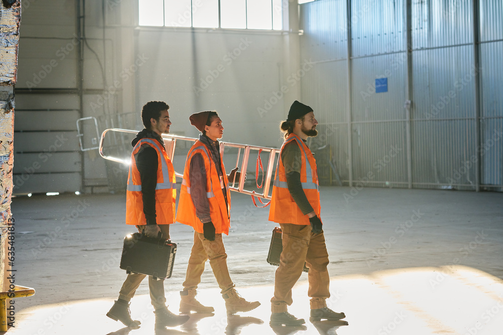 Poster side view of three young engineers or technicians of warehouse carrying stepladder and toolboxes whi