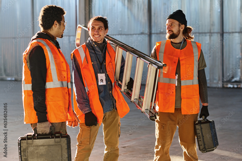 Leinwandbilder Three young intercultural men in protective fluorescent jackets discussing working points while one of them explaining something to colleagues