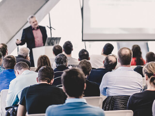 Obraz premium Man giving presentation in lecture hall. Male speeker having talk at public event. Participants listening to lecture. Rear view, focus on people in audience.