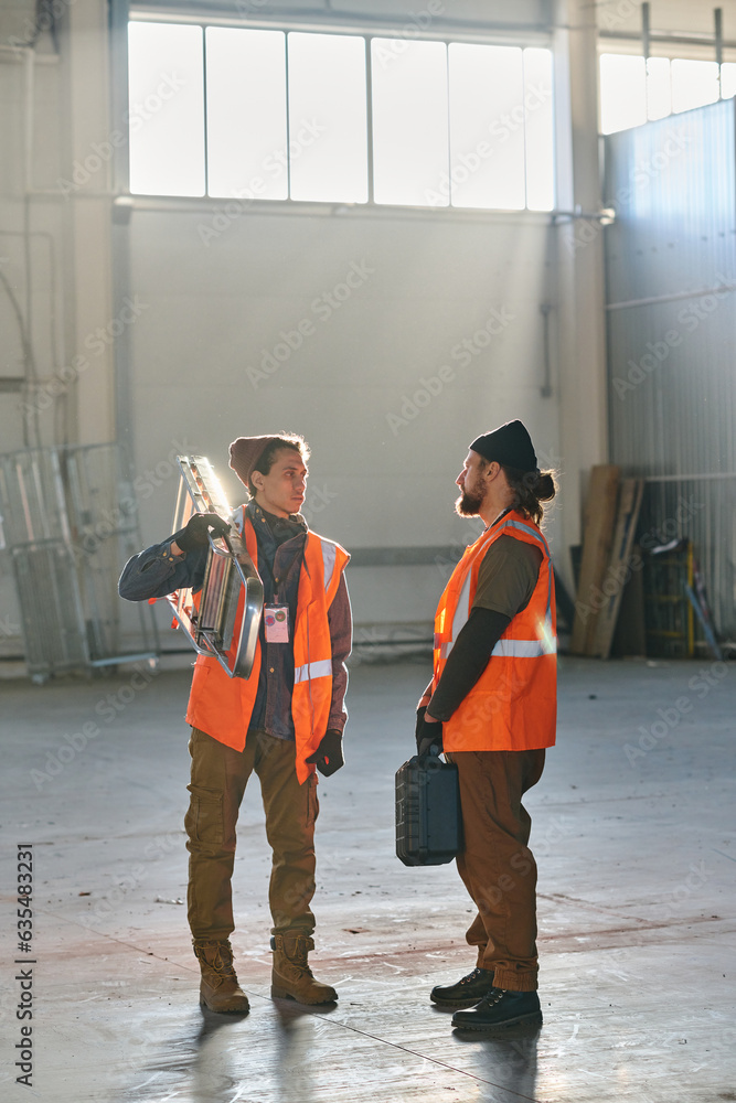 Poster two young engineers in protective reflective jackets discussing something at working meeting while o