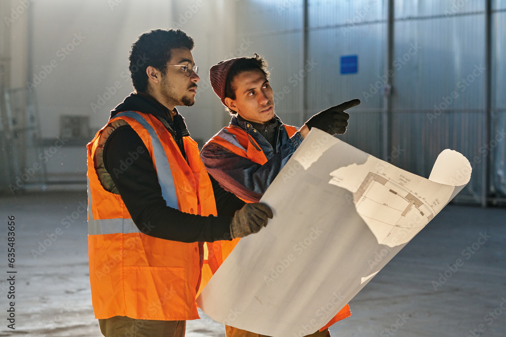 Leinwandbilder One of two engineers pointing forwards while explaining points of sketch to his colleague holding big blueprint at working meeting in warehouse