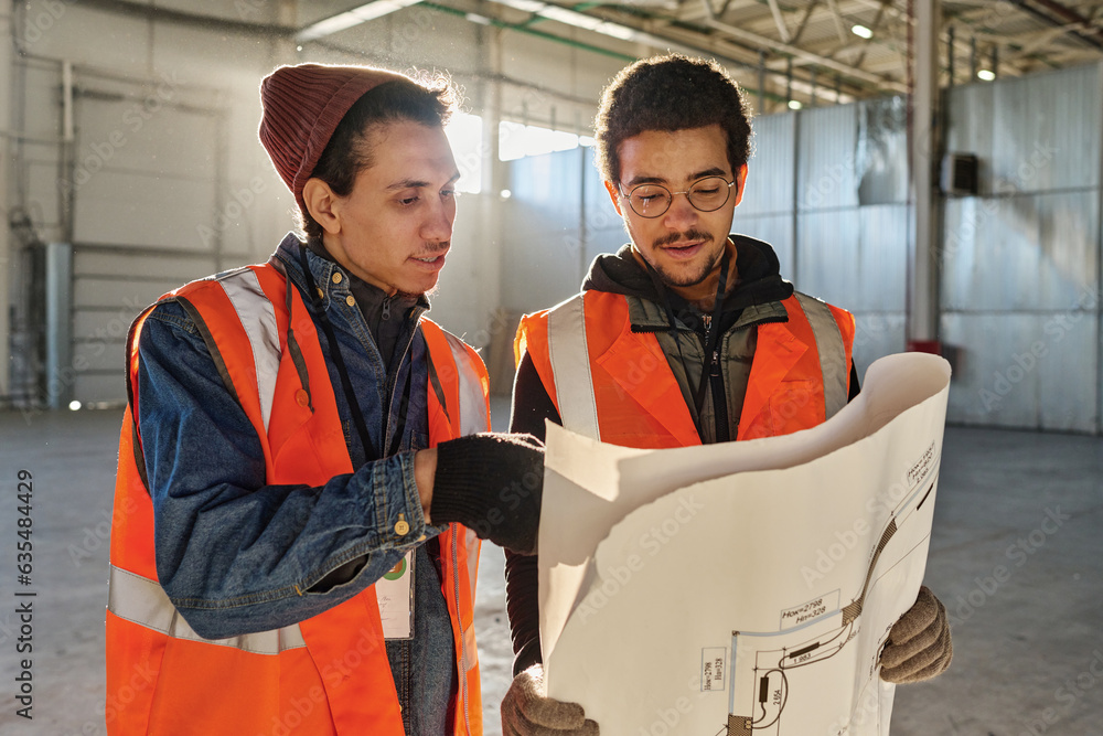 Poster two young multiethnic engineers looking at building plan on blueprint while one of them pointing at 