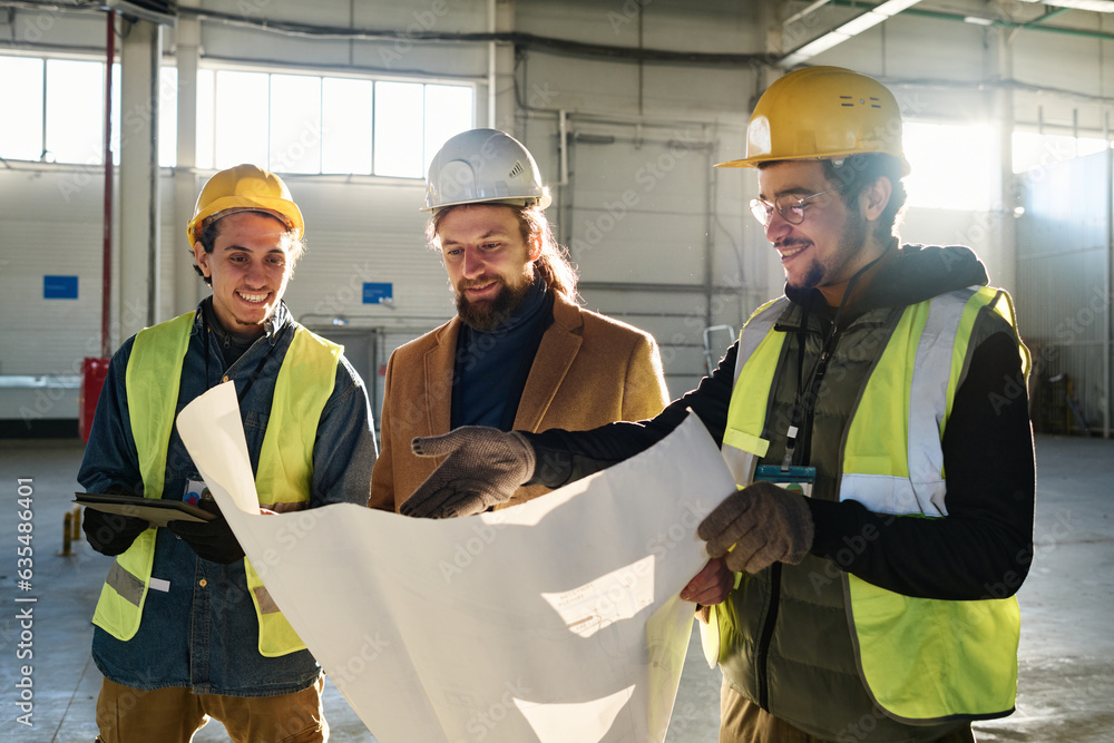 Leinwandbilder Young smiling engineer pointing at building plan in blueprint while explaining it to contractor and colleague at meeting in warehouse