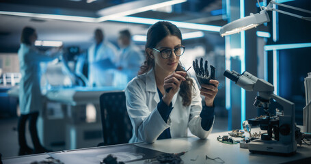 Female Engineer Fixing a High-Tech Bionic Hand Component. On Blurred Background Industrial Robotics Specialists in Lab Coats Working on a Mobile AI Robot in a Tech Factory Facility - obrazy, fototapety, plakaty