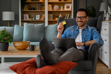 Happy businessman with credit card and laptop doing home shopping while relaxing on armchair
