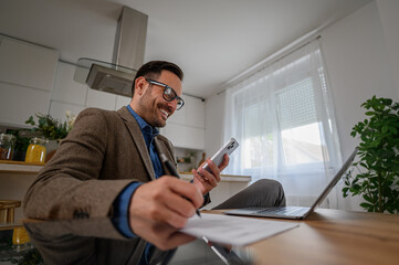 Fototapeta premium Low angle view of smiling businessman in suit using smart phone with laptop on desk at home office