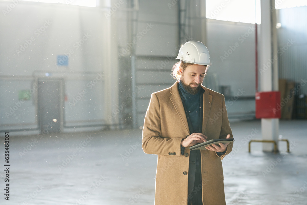 Poster Young bearded builder or solopreneur in hardhat scrolling in tablet while standing in front of camera in spacious sunlit warehouse and networking
