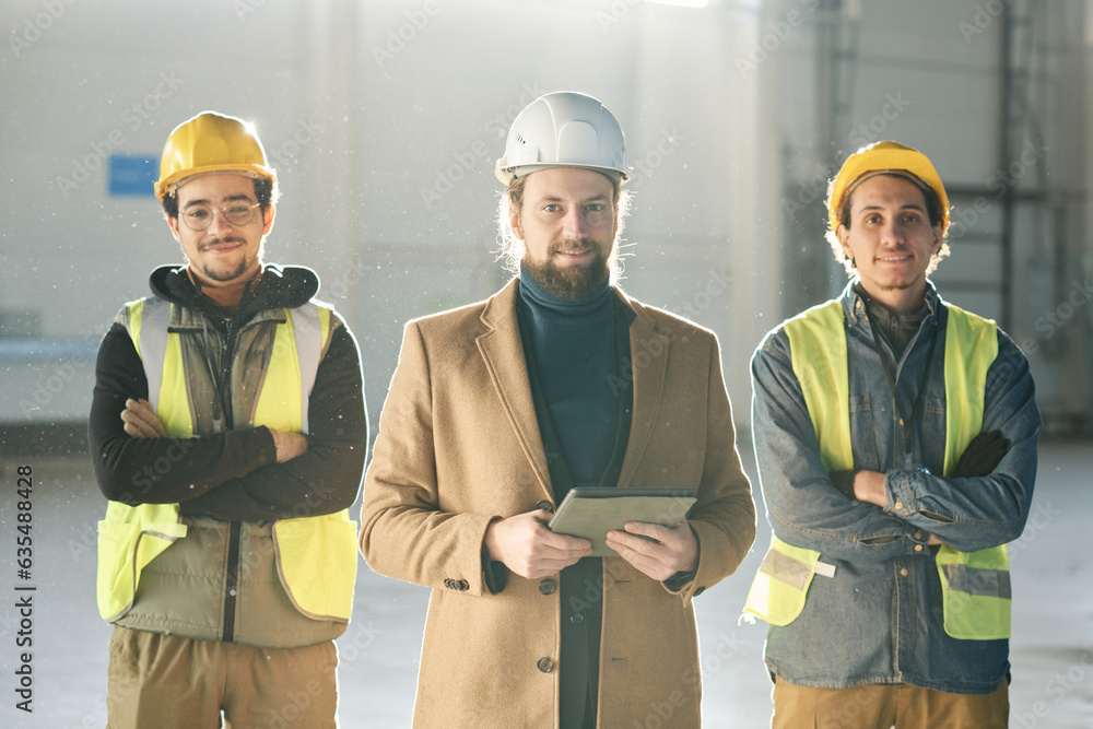 Leinwandbilder Young successful foreman with tablet standing between two engineers in hardhats and protective fluorescent vests and looking at camera