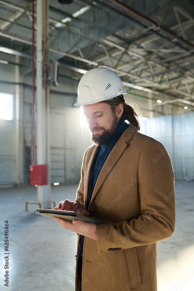 Poster young serious contractor looking through online information on tablet screen while standing in front