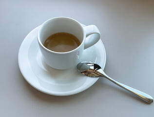 A cup of espresso sits on a white plate with a silver spoon sitting besides it. 