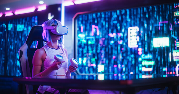 Cyberpunk Gamer Girl Wearing a Virtual Reality Headset and Using Controllers in a Neon Futuristic Room. Young Cosplay Female Exploring VR Metaverse Online While Sitting Behind a Desk