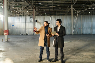 Long shot of two young intercultural entrepreneurs in formalwear looking around area for new office while standing in spacious warehouse