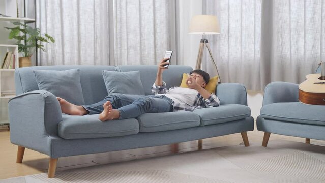Full Body Of Asian Teen Boy Using Smartphone Taking Selfie While Lying On Sofa In The Living Room At Home. Smiling, Showing Peace, Thumbs Up, And Heart Gesture
