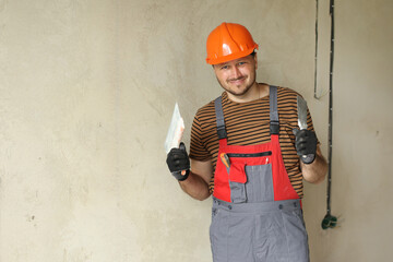 Smiling repairman in overalls and protective orange helmet holds two spatulas for plastering...