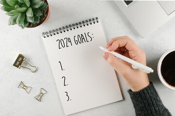 Woman's hand writing 2024 Goals in notebook. Laptop, home plant, cup of coffee on light table. Top view, flat lay - 635495266