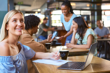 Woman Working On Laptop In Foreground With Couple Meeting Socially Behind In Busy Coffee Shop