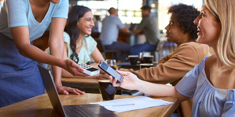 Woman Using Laptop In Coffee Shop Paying Bill With Contactless Mobile Phone Payment