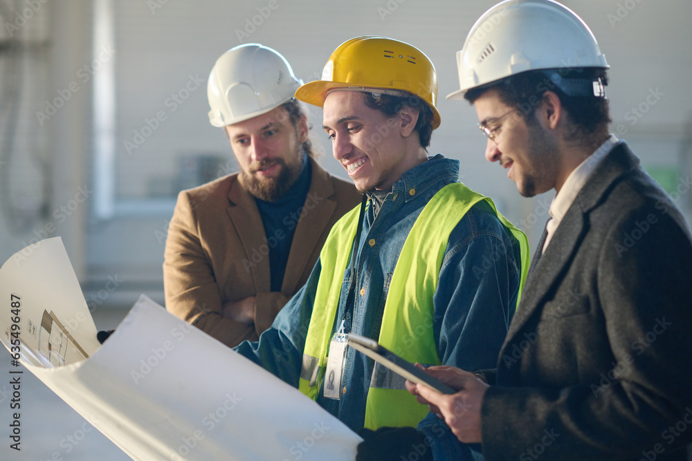 Aufkleber focus on young smiling engineer in workwear and hardhat holding blueprint with building plan while s