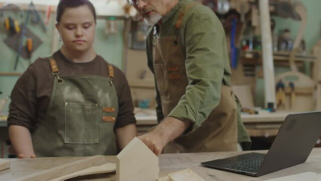 Confident mature master talking to young female apprentice with Down syndrome during presentation of new wooden details for restoration of furniture while showing her some of them on laptop screen