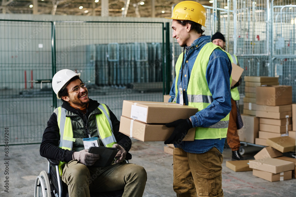 Poster Happy young engineer in wheelchair looking at colleague carrying stack of packed cardboard boxes while talking to him during work in warehouse