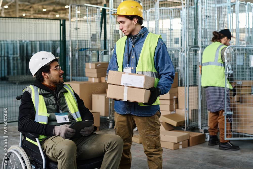 Poster young engineer with stack of packed cardboard boxes talking to colleague sitting in wheelchair and u