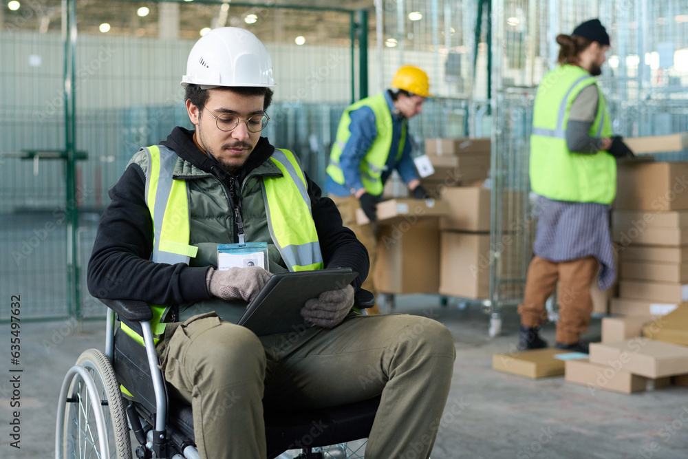 Poster young serious worker of warehouse sitting in wheelchair in front of camera against two colleagues lo