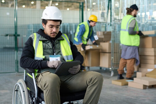 Young serious worker of warehouse sitting in wheelchair in front of camera against two colleagues loading boxes on carts and using tablet