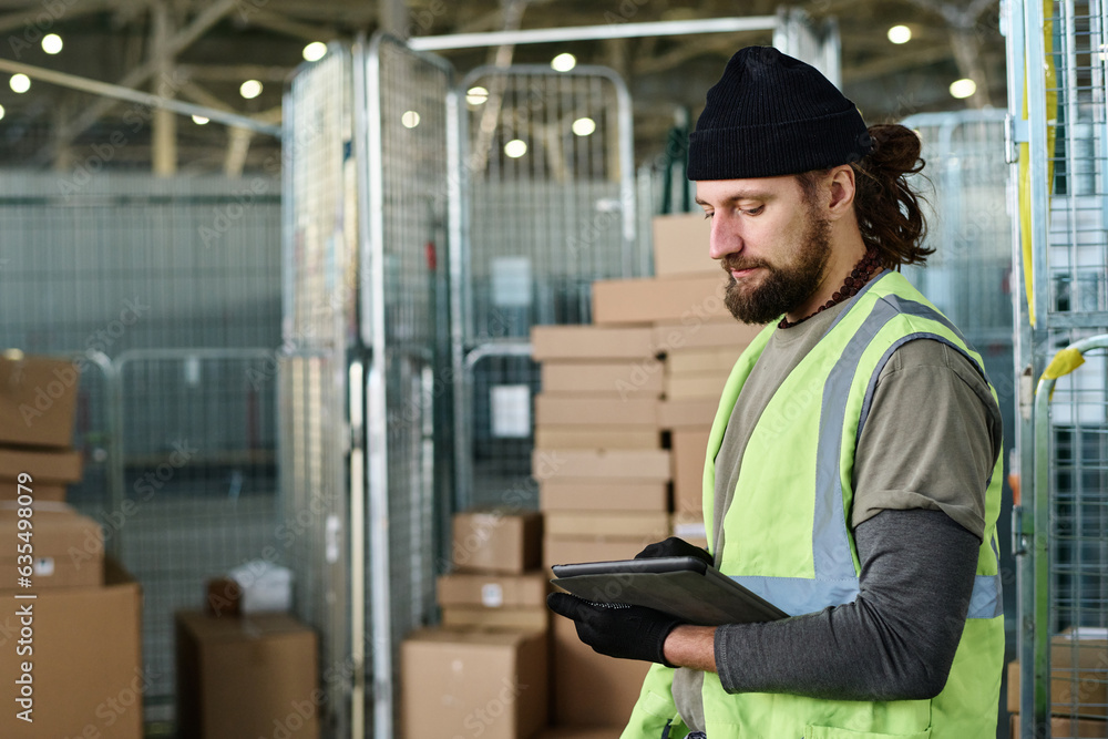 Poster side view of bearded worker of warehouse in reflective vest looking through database in tablet while