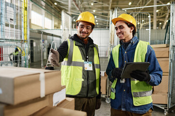 Happy young man in workwear using tablet and listening to colleague pointing at stack of packed...