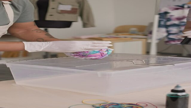 Vertical cropped shot of team of female fashion designers using vibrant fabric dyes while creating tie dye print on white t-shirt in bright workshop