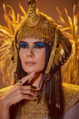 Elegant woman in egyptian look and headdress posing near blurred plants on brown background